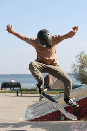 Image of boy doing an Ollie on a skateboard. he is looking down. Wearing an orange shite and green-grey pants. It is a sunny day in a skate park in Orillia, Ontario