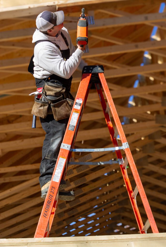 Adult and teenager standing outdoors at a construction site, smiling at the camera, with the teen wearing a safety helmet and college hoodie and the adult wearing sunglasses and a plaid shirt.