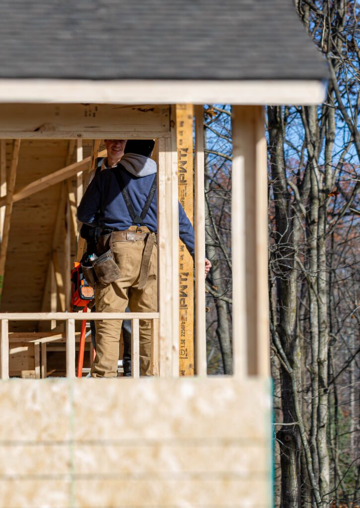 Construction worker seen from behind inside a partially built wooden structure, working on framing while trees are visible in the background.