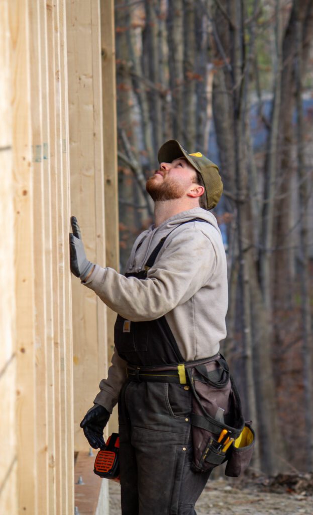Construction worker wearing gloves and a tool belt inspects a newly framed wooden wall at an outdoor building site surrounded by trees.